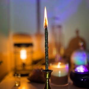 May include: A burning dark green candle in a brass candlestick. The candle has a textured surface and a bright orange and yellow flame. The candlestick has a round base and a flared top. The background is blurred, with other candles and objects.