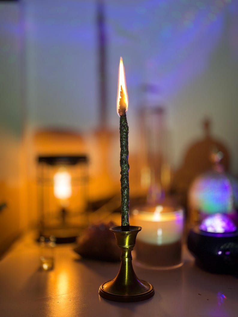 May include: A burning dark green candle in a brass candlestick. The candle has a textured surface and a bright orange and yellow flame. The candlestick has a round base and a flared top. The background is blurred, with other candles and objects.