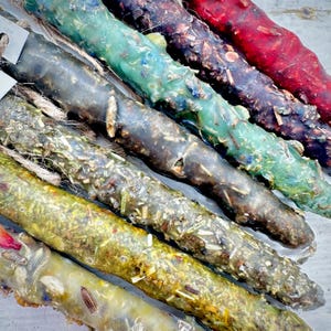 May include: A close-up of several colorful, handmade candles. The candles are cylindrical and decorated with dried herbs and flowers. Colors include green, brown, red, and yellow. Each candle has a natural, rustic appearance and is tied with twine.