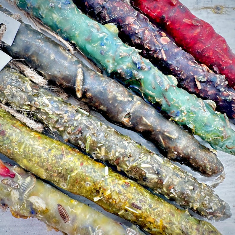 May include: A close-up of several colorful, handmade candles. The candles are cylindrical and decorated with dried herbs and flowers. Colors include green, brown, red, and yellow. Each candle has a natural, rustic appearance and is tied with twine.