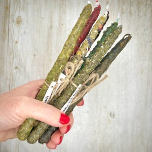 May include: A bundle of seven herbal candles in varying shades of green, brown, and red, held together with twine. Some candles have visible herbs and flowers. Small white tags are attached to the twine. The text "Ease The Ties" is visible on one tag.