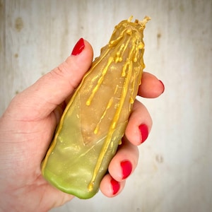 May include: A handmade candle with a unique design. The candle is a combination of green and gold colors, with gold drips cascading down the sides. The candle is held in a hand with red nail polish.