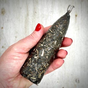 May include: A cylindrical candle in a dark charcoal gray color, featuring a textured surface embedded with numerous small, light-colored dried botanical pieces and herbs throughout the wax, topped with a single visible cotton wick at the point.