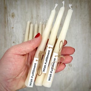 May include: A set of off-white taper candles, each featuring a small white rectangular tag tied with brown twine. The tags are printed with the text Wish Fulfillment. The candles are slender with a smooth, matte finish and a simple wick.