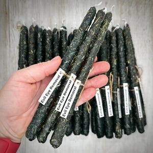 Puede incluir: Una colección de velas verde oscuro, infundidas con hierbas, algunas sostenidas en una mano. Cada vela está etiquetada con "Evil Eye Removal". Las velas son cilíndricas y parecen hechas a mano, con una estética rústica.