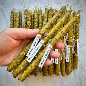 Può includere: Un fascio di candele affusolate verdi e strutturate. Ogni candela è adornata da un'etichetta bianca con la scritta "Attract Clients". Le candele sono legate con spago.