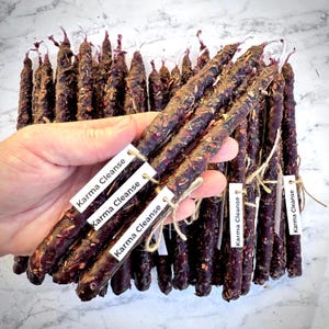 May include: A collection of dark purple ritual candles, adorned with dried herbs. Each candle has a white tag that reads "Karma Cleanse" tied with twine. The candles are cylindrical and appear to be hand-rolled.