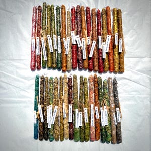 May include: A collection of forty long, slender, textured candles arranged in two rows. Each candle features a unique color and is tied with a small white paper tag displaying text such as Abundance, Protection, Love, Discipline, and New Moon.