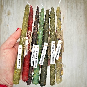 May include: A collection of eight colorful, handmade spell candles. Each candle is a different color, including green, red, and brown, and is adorned with herbs and twine. Small white tags with phrases like "Open Roads" and "Forgiveness" are attached.