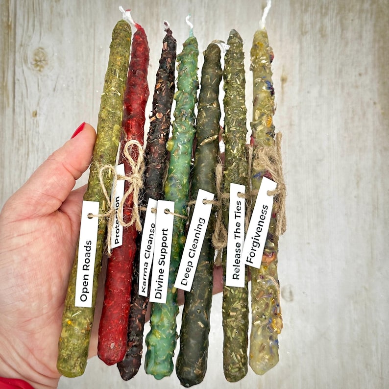 May include: A collection of eight colorful, handmade spell candles. Each candle is a different color, including green, red, and brown, and is adorned with herbs and twine. Small white tags with phrases like "Open Roads" and "Forgiveness" are attached.