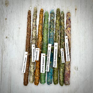 May include: A collection of ten tall, textured candles in various colors, including brown, green, and blue, with attached tags. The tags read "Ancestral Power," "Forgiveness," "Gratitude," "Divine Support," "After Conflict Cleanse," "Health Restoration," "Stress Relief," and "Energy Restoration."