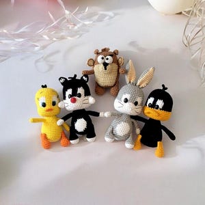 May include: A collection of five handmade crochet cartoon character dolls, including a yellow Tweety Bird, a black and white Sylvester the Cat, a brown Taz, a gray Bugs Bunny, and a black Daffy Duck. The dolls are arranged on a white surface with balloons in the background.