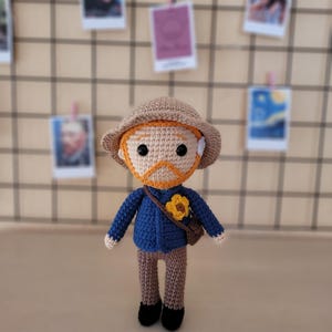 Könnte beinhalten: Gehäkelte Puppe eines Mannes mit orangefarbenem Haar und Bart, der eine blaue Jacke, braune Hose und einen beigefarbenen Hut trägt. Die Puppe hat eine gelbe Blume auf der Jacke und eine braune Umhängetasche. Schwarze Schuhe vervollständigen das Outfit.