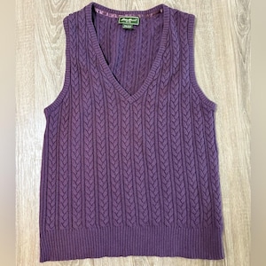 May include: A purple sleeveless sweater vest featuring a vertical cable knit pattern, a V-neckline, and a ribbed hem. The interior neck label displays the text Eddie Bauer, with additional text indicating the size and material composition.