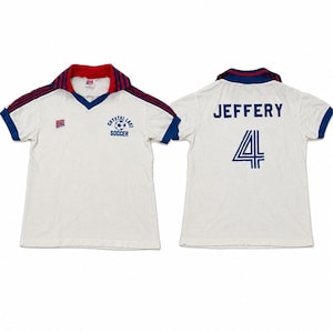 May include: A white short-sleeved football shirt with a red and blue collar, blue sleeve cuffs, and red and blue stripes on the shoulders. The front features a Crystal Lake Soccer logo and a small patch. The back displays the name JEFFERY and number 4.