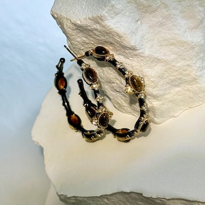18K gold tone bamboo hoop earrings with tiger’s eye stones and rhinestones, standing upright and leaning against stone prop to highlight textured gold surface and gemstone shine