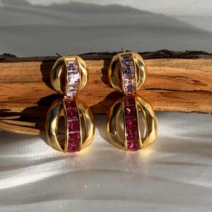 May include: A pair of gold-toned earrings with a geometric design. Each earring features two sections with a series of pink and red rectangular stones. The earrings have a textured, rounded shape and are displayed on a wooden surface.
