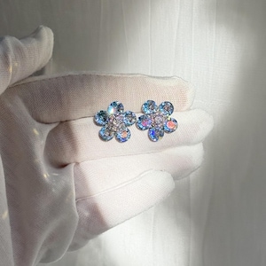 May include: A pair of flower-shaped stud earrings. Each earring features a blue and iridescent petal design surrounding a cluster of clear crystals. The earrings are held in a white gloved hand.