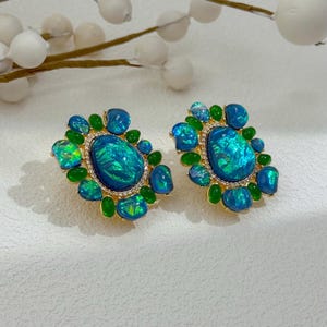 May include: A pair of ornate stud earrings featuring large, oval, iridescent blue-green stones surrounded by smaller blue and green stones. The earrings have a gold-colored setting and are accented with small, clear crystals.
