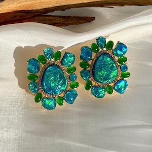 May include: A pair of gold-toned earrings featuring large, oval-shaped, iridescent teal-green stones surrounded by smaller teal and green stones. The earrings have a halo of small, clear stones around the central stone and are set against a white background.