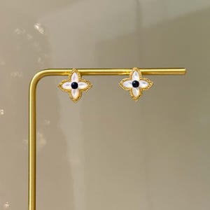Peut inclure: Une paire de boucles d'oreilles à clous dorées. Chaque boucle d'oreille présente un motif de fleur à quatre pétales avec un centre blanc et un point noir au milieu. Les boucles d'oreilles sont exposées sur un support doré.