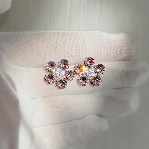 Pode incluir: Dois brincos de botão em forma de flor com cristais rosa claro e transparentes brilhantes. Os brincos são exibidos em um fundo branco, destacando seu design intrincado e brilho. O design floral adiciona um toque de elegância.