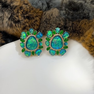 May include: A pair of ornate stud earrings. Each earring features a large, iridescent, oval-shaped center stone in shades of teal and green, surrounded by smaller green stones and a gold-colored setting. The earrings are displayed on a white surface.