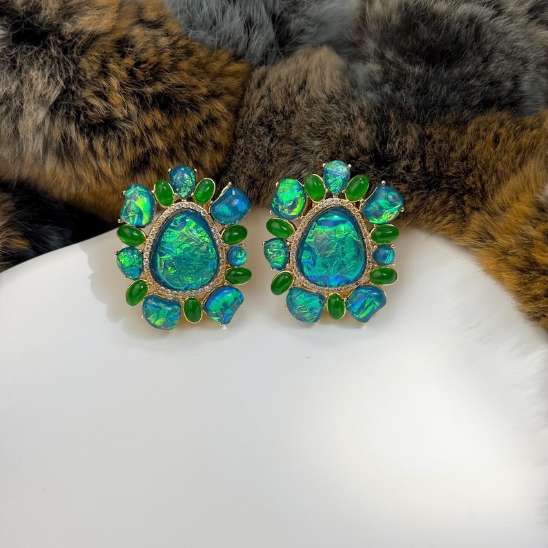 May include: A pair of ornate stud earrings. Each earring features a large, iridescent, oval-shaped center stone in shades of teal and green, surrounded by smaller green stones and a gold-colored setting. The earrings are displayed on a white surface.