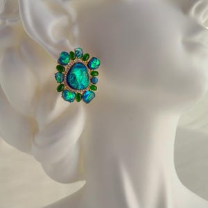 May include: A gold-toned stud earring featuring a large, iridescent teal stone surrounded by smaller green and teal stones. The central stone has a textured appearance, and the setting includes small, clear accent stones.