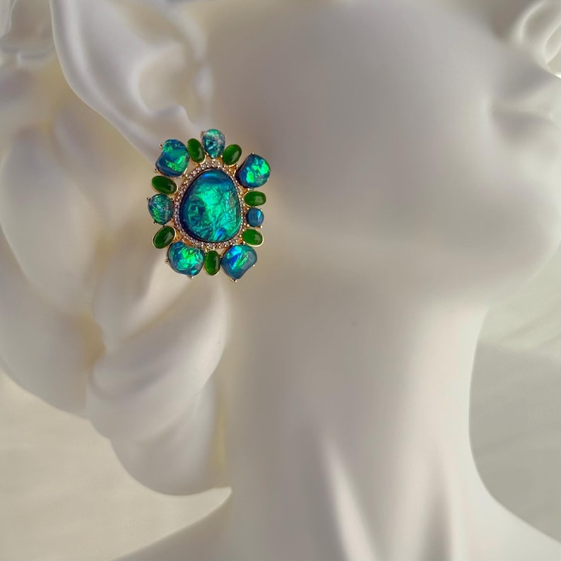 May include: A gold-toned stud earring featuring a large, iridescent teal stone surrounded by smaller green and teal stones. The central stone has a textured appearance, and the setting includes small, clear accent stones.