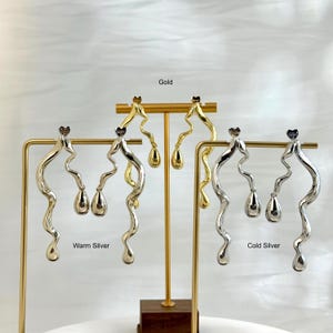Puede incluir: Pendientes en tonos dorados y plateados expuestos en soportes. Los pendientes tienen un diseño ondulado con un colgante en forma de lágrima. Los textos "Gold", "Warm Silver" y "Cold Silver" son visibles.