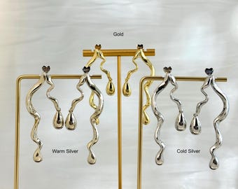May include: Earrings in gold and silver tones are displayed on stands. The earrings have a wavy design with a teardrop-shaped dangle. The text "Gold", "Warm Silver", and "Cold Silver" are visible.