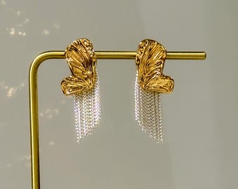 May include: A pair of gold-colored earrings with a textured, leaf-like design. Each earring features a cascade of silver-colored chain fringe, creating a layered effect. The earrings are displayed on a gold-colored stand.