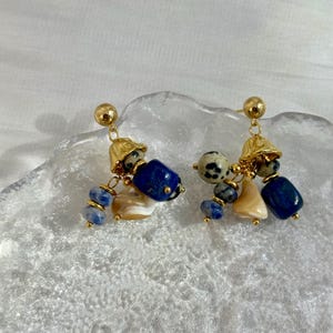May include: A pair of gold-toned dangle earrings. Each earring features a gold bell-shaped top, a dark blue stone, a speckled stone, and smaller blue and tan stones. The earrings are displayed on a textured surface.