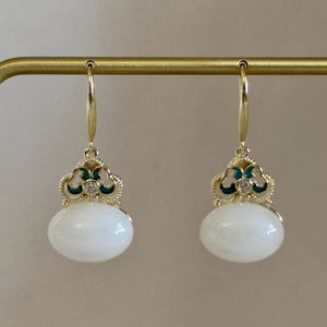 May include: A pair of gold-toned drop earrings. Each earring features a white, oval-shaped stone suspended from a decorative top with green and gold accents. A small clear gem is set in the center of the top.