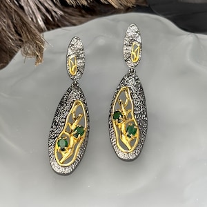 May include: A pair of dangle earrings with a textured, dark gray metal finish. The earrings feature gold-colored filigree designs with three small, green gemstones. The earrings have a teardrop shape and a post-back closure.