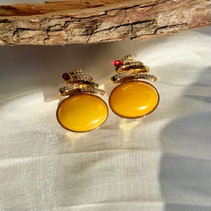 Può includere: Un paio di orecchini color oro con grandi pietre gialle ovali. Ogni orecchino ha un design a spirale con accenti chiari e rossi. Gli orecchini sono posti su uno sfondo bianco con un elemento in legno.