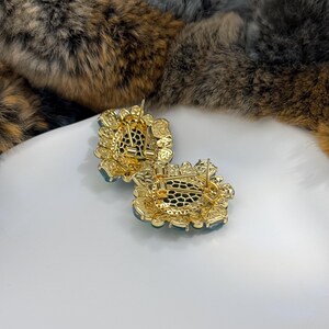 May include: A pair of gold-toned earrings with a floral design. Each earring features a central teal-colored stone surrounded by smaller clear stones. The earrings have a post-back closure and are set on a white surface.