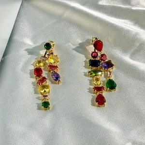 May include: A pair of gold-toned earrings with a cascade of colorful gemstones. The earrings feature a variety of shapes and colors, including red, yellow, green, blue, and purple. The gemstones are arranged in a linear, dangling design.