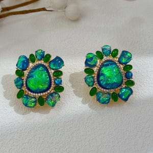 May include: A pair of square-shaped earrings with a gold-colored setting. Each earring features a large, oval-shaped, iridescent green and blue stone surrounded by smaller blue and green stones and green teardrop-shaped stones. The center stone is framed by small, clear crystals.