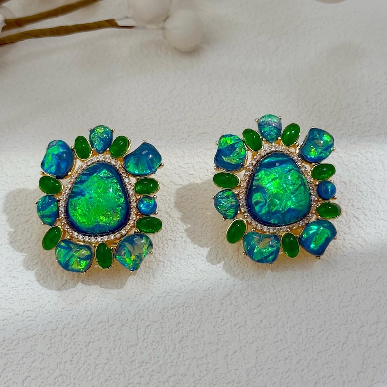 May include: A pair of square-shaped earrings with a gold-colored setting. Each earring features a large, oval-shaped, iridescent green and blue stone surrounded by smaller blue and green stones and green teardrop-shaped stones. The center stone is framed by small, clear crystals.