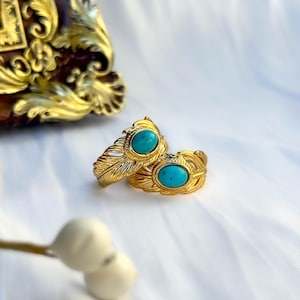 May include: Two gold-toned rings featuring a detailed feather-like texture along the band. Each ring is set with a single, smooth, oval-shaped turquoise stone in the center. The metal has a polished finish, highlighting the intricate grooves.