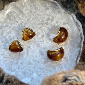 May include: Four earrings with a gold trim. Two are triangular, and two are semi-circular. The earrings have a marbled, amber-colored resin center. The earrings are displayed on a textured, white surface.