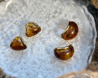 Könnte beinhalten: Vier Ohrringe mit goldfarbenem Rand. Zwei sind dreieckig und zwei halbkreisförmig. Die Ohrringe haben einen marmorierten, bernsteinfarbenen Kunstharzmittelteil. Die Ohrringe werden auf einer strukturierten, weißen Oberfläche präsentiert.