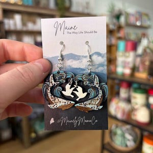 May include: Wooden earrings featuring a wave design with a white bird silhouette. The earrings are on a card with the text "Maine The Way Life Should Be" and the shop's name. The earrings are held up by silver hooks.