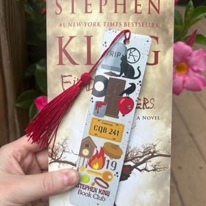 May include: A Stephen King novel with a decorative bookmark. The bookmark features a red tassel and various illustrations, including a tombstone, black cat, door, apple, and the text "CQB 241". The book's title is "Stephen King".