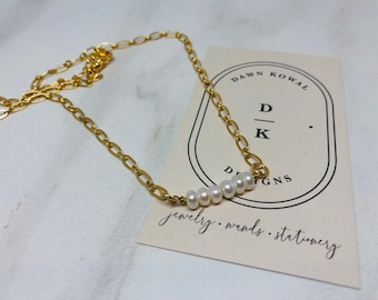May include: A gold chain necklace with a row of small white pearls. The necklace is on a white surface with a business card that reads "Dawn Kowal D/K Designs jewelry. wands. stationery."