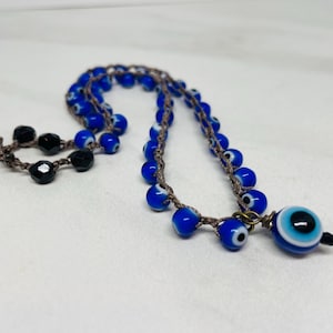 May include: A necklace featuring blue glass beads with an evil eye design, strung on a brown cord. The necklace also includes black beads and a larger evil eye pendant. The jewelry is displayed on a white surface.
