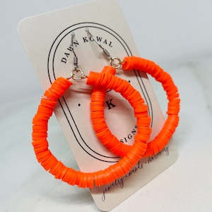 May include: Bright orange hoop earrings made of small, flat, circular beads. The earrings are displayed on a white card with the text "DAWN KOWAL" and other text. The earrings have silver-colored hooks.
