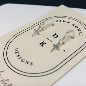 May include: A pair of small, silver hoop earrings with a cross charm. The earrings are displayed on a cream-colored card with the text "DAWN KOWAL DESIGNS" and the initials "DK". The earrings are simple and elegant.